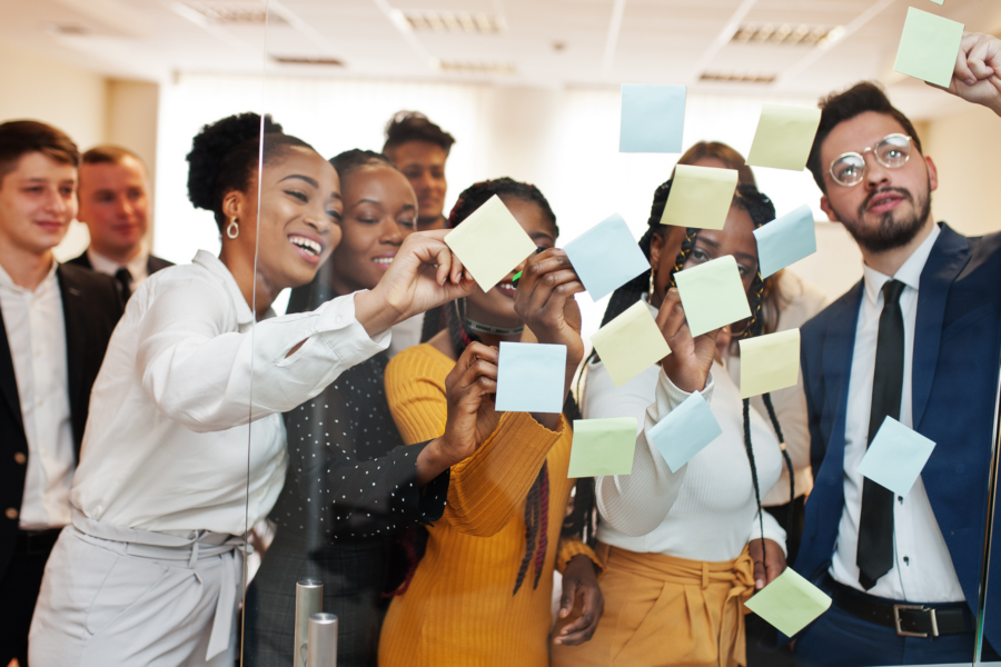 A group of office people smile and put up post-its on a trasnparent screen.