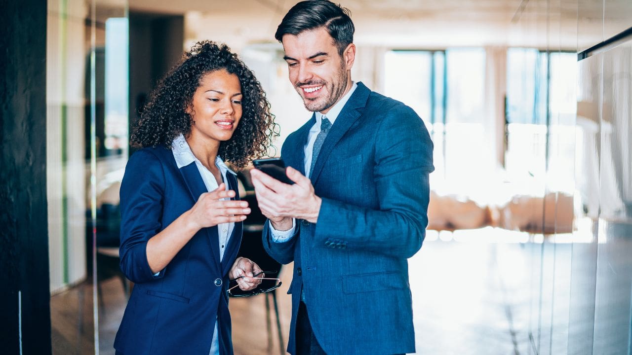 A happy business woman and business man, both in blue suits, looking at a phone in a bright office.