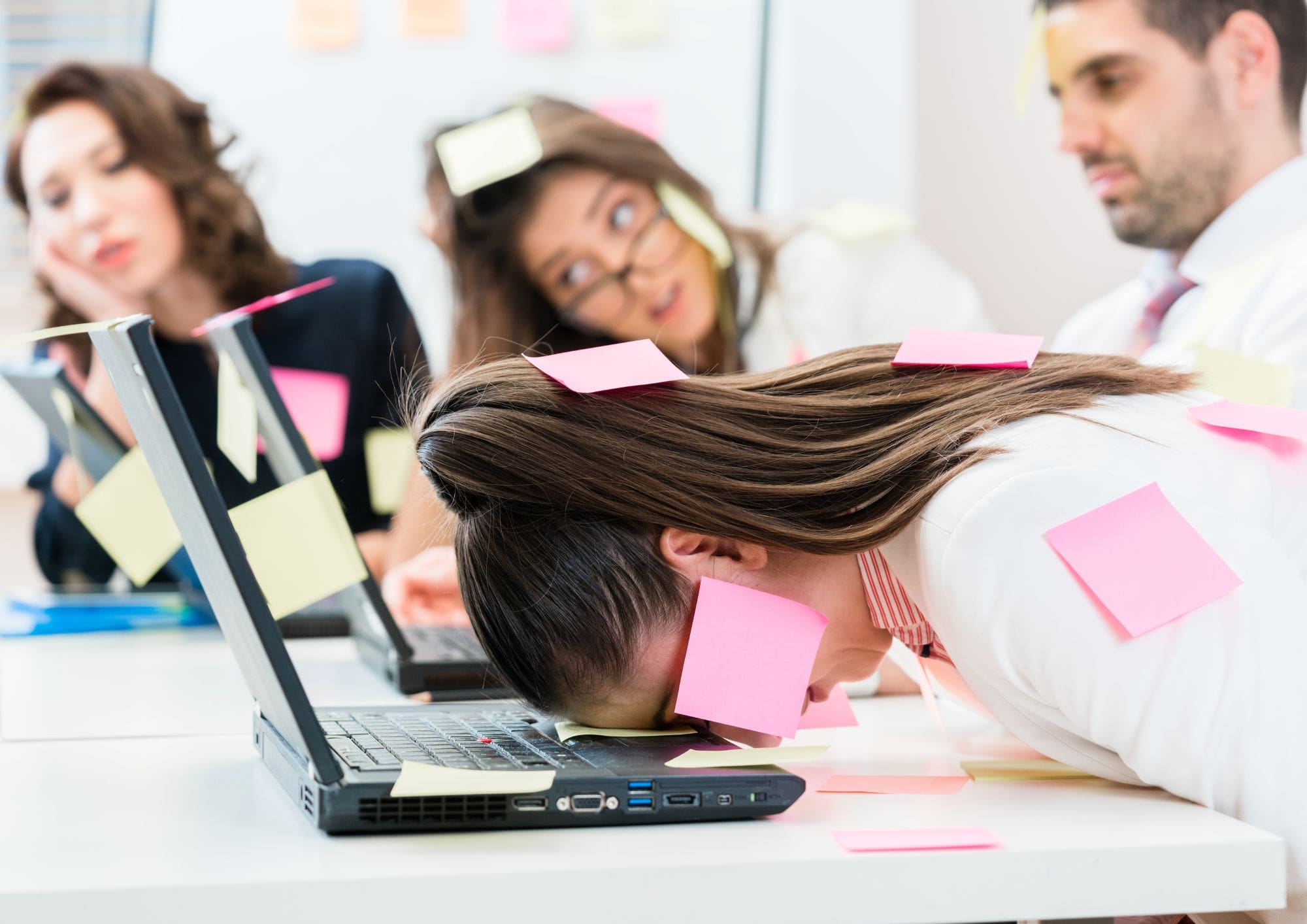 4 extremely stressed out business people, with a business woman covered in post-it notes and her head on the laptop on the desk.