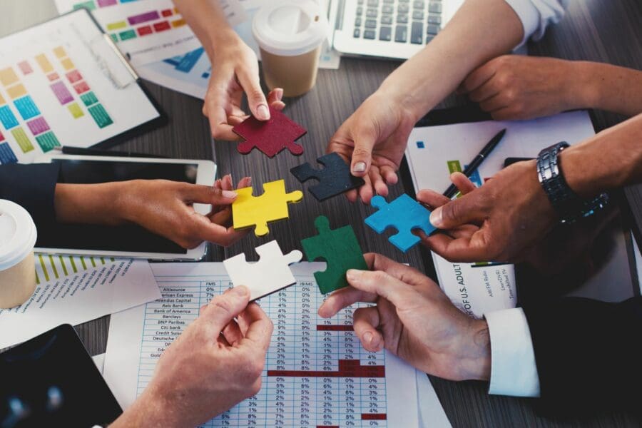 A messy meeting table with reports and coffee cups, etc. There are Multiple arms with hands all meeting in the middle. Each hand holds a different coloured jigsaw puzzle piece.