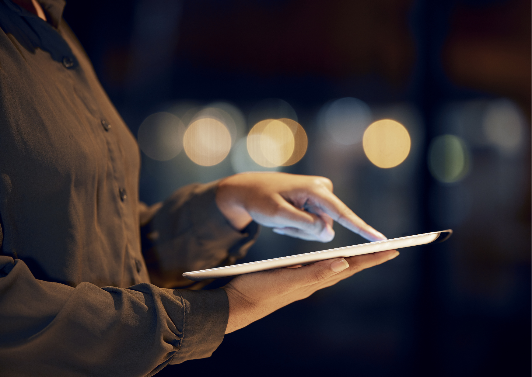 A woman in a brown shirt uses an ipad. There's a window in the back, showing it is dark outside.