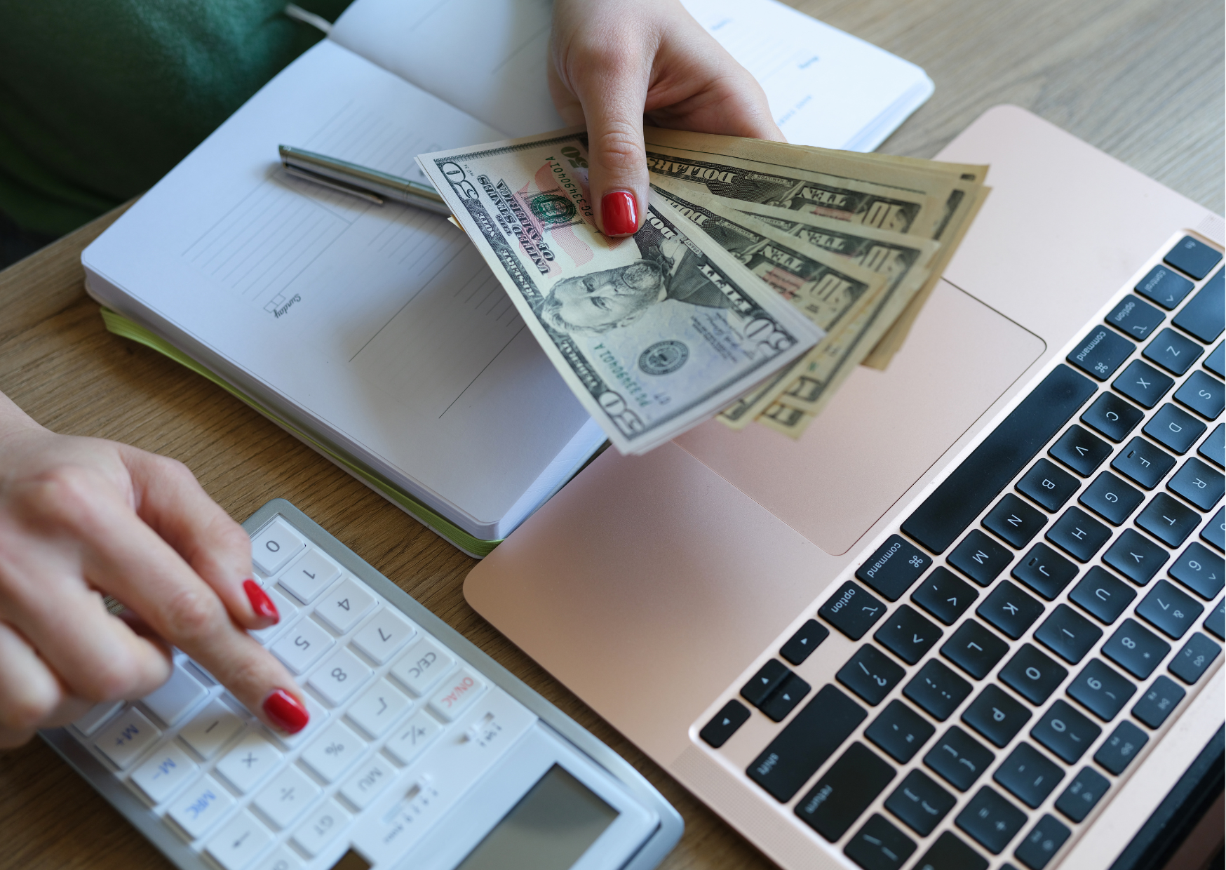 A woman's hands holding money and using a calculator. There's a laptop, notebook and pencil under her hands.