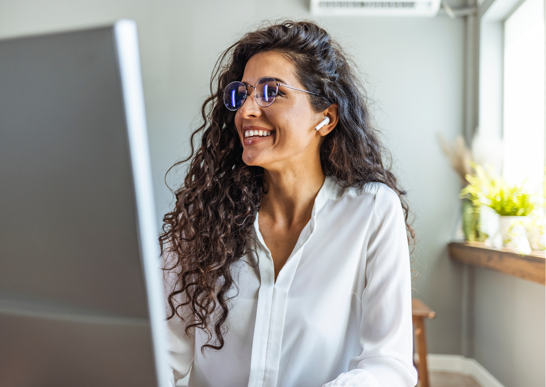 A long-haired woman with glasses in an office smiles at the computer screen.