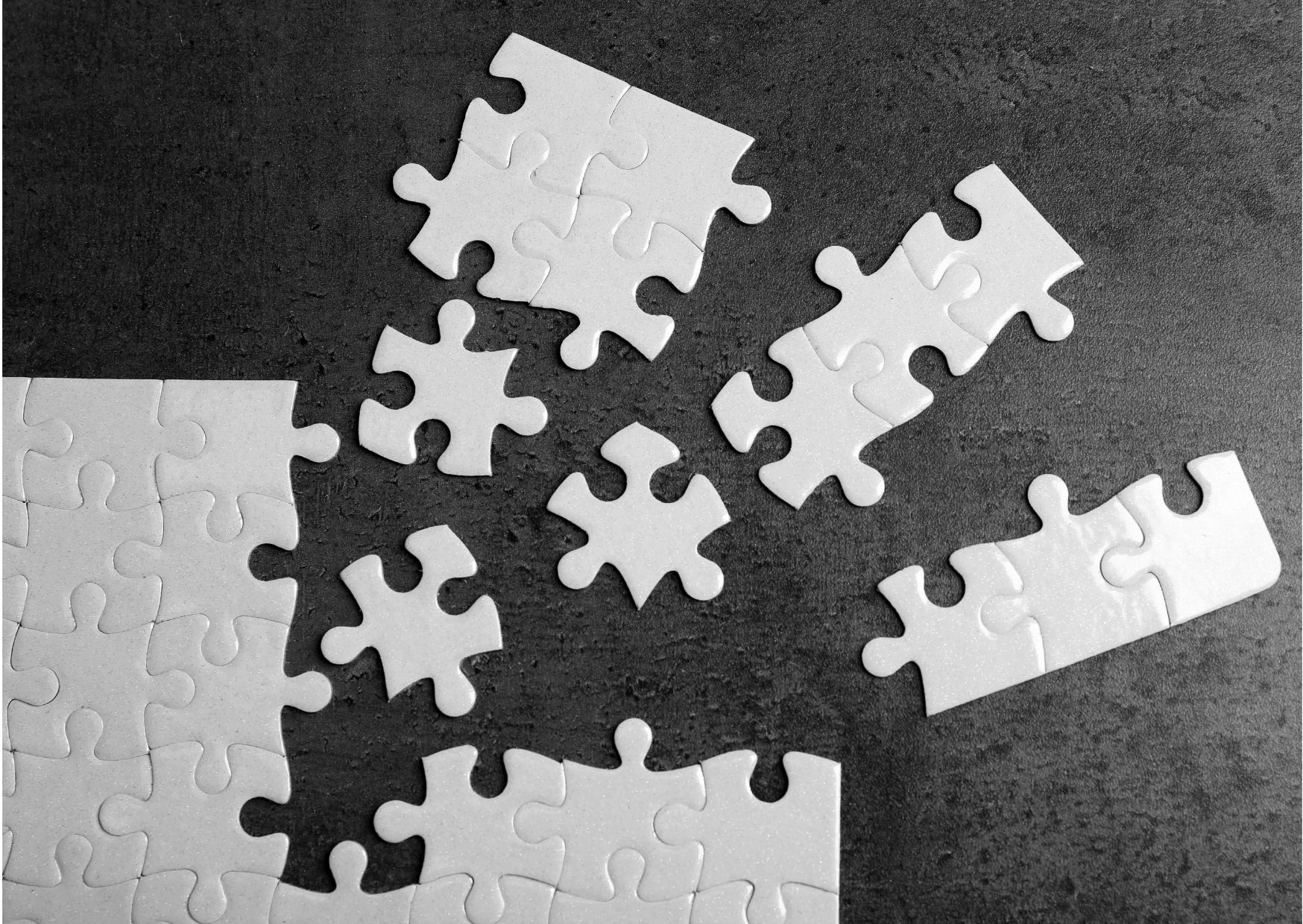 On a dark table are white puzzle pieces. Some are joined, some brand off.