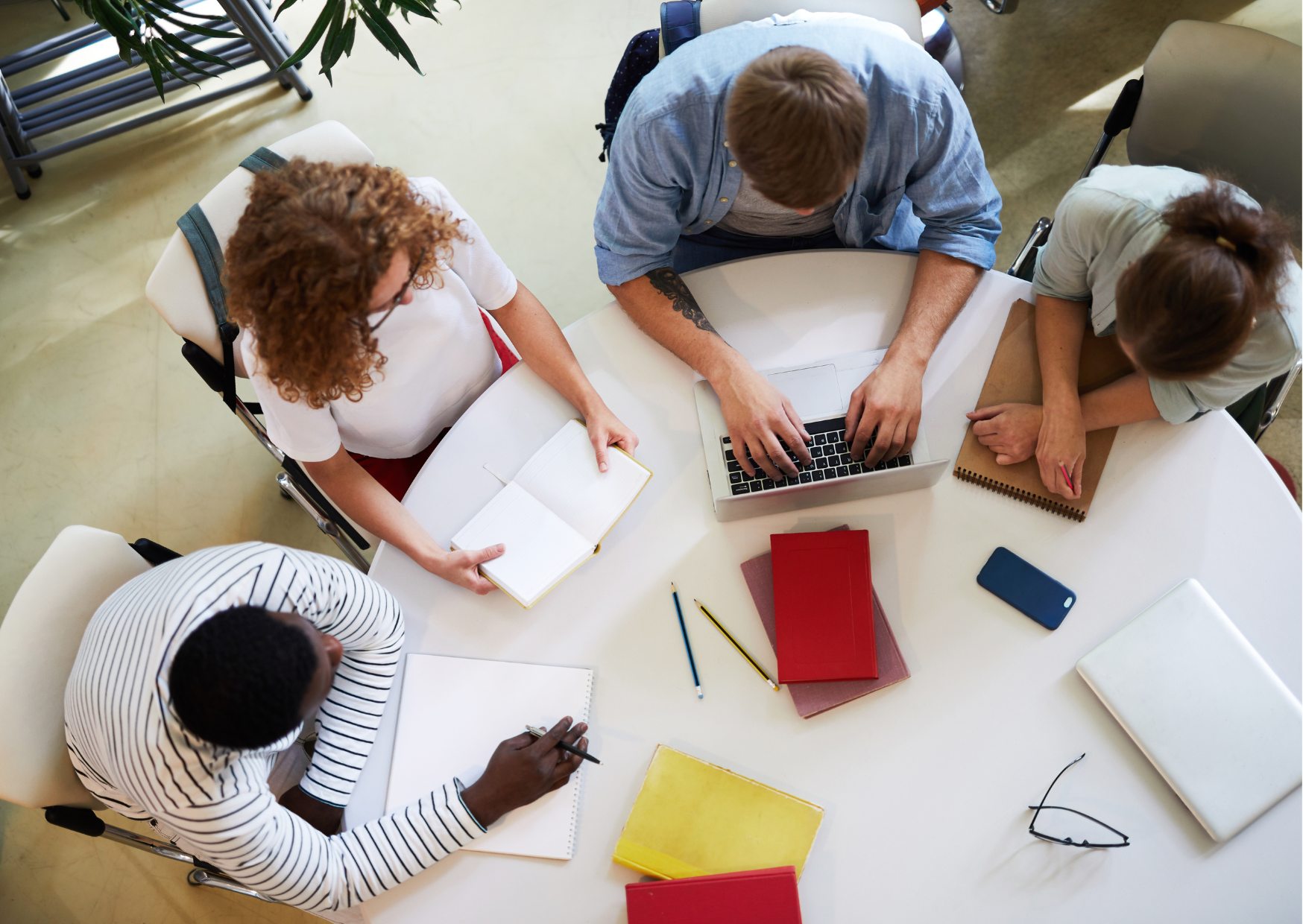 An aerial view of 4 people sitting at a white office desk with pens, paper, laptop, glasses, etc.