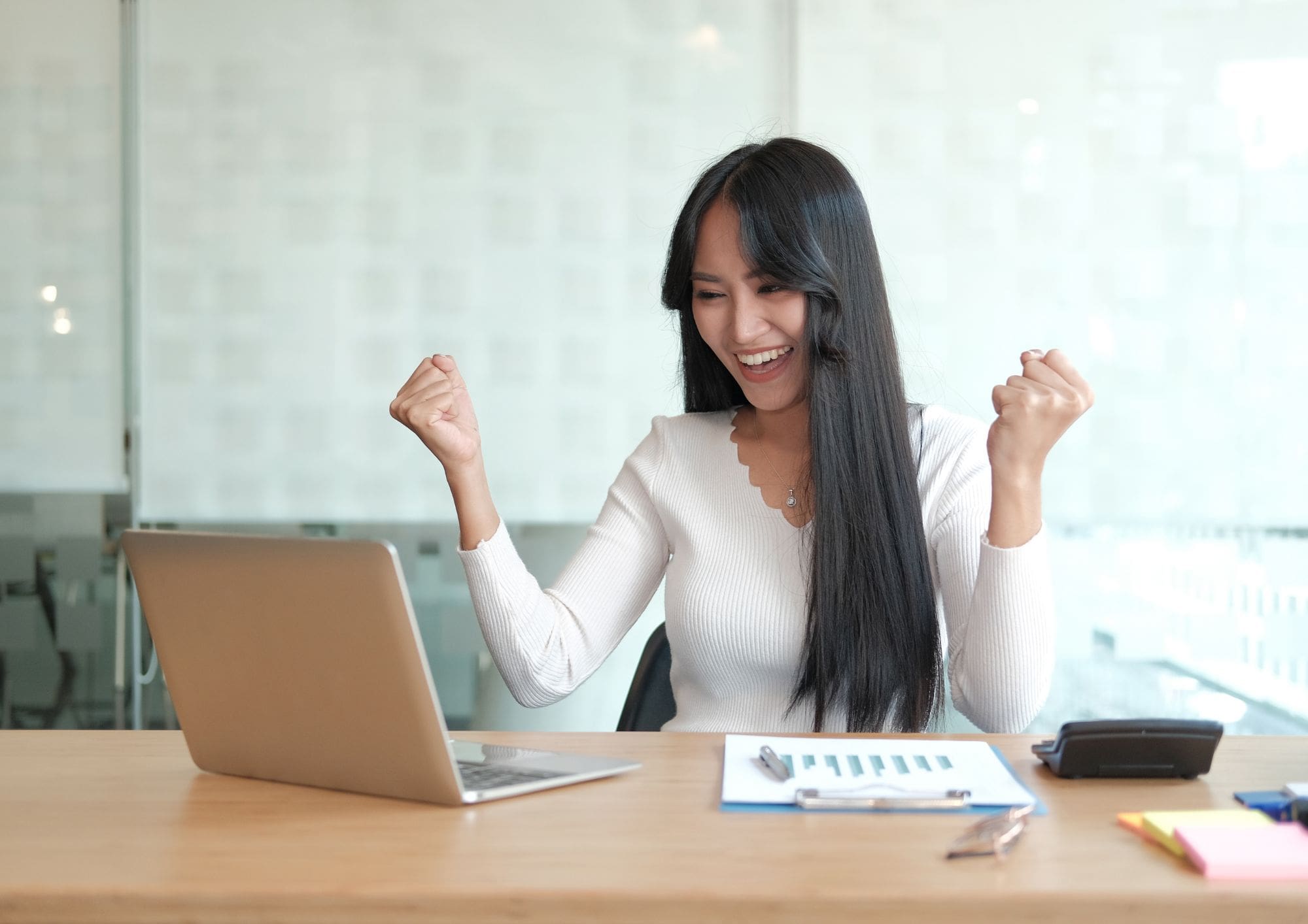 A businesswoman in an office celebrating a win in front of a laptop.